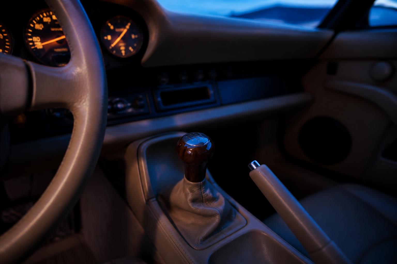 A man's hand resting on the wood-and-leather gear shifter of a Porsche 911, Mojave Beige interior, predawn cobalt blue light coming through the windshield