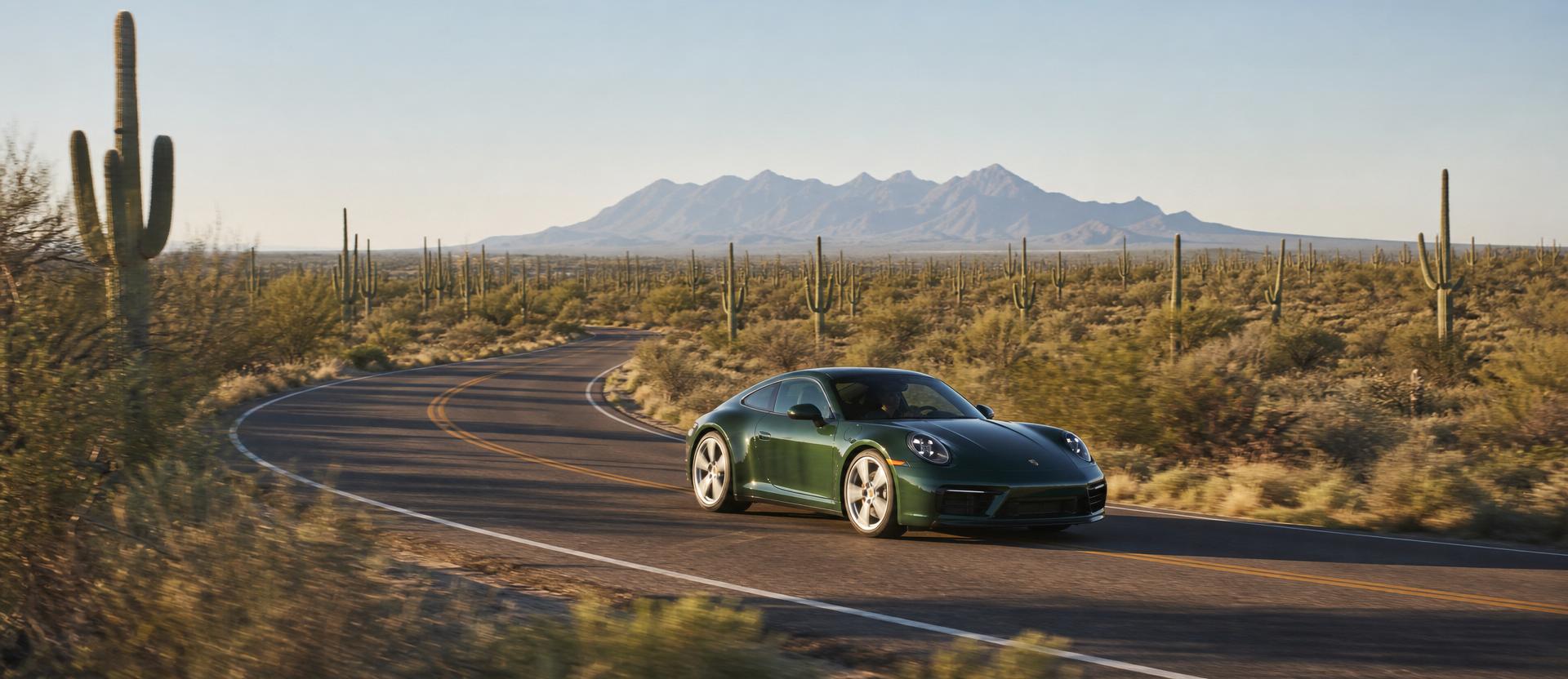 An Aventurine Green Porsche 911 in motion mid-corner on the empty two-lane ribbon of Rio Verde Road, a saguaro flat stretching out around it, the blue silhouette of Four Peaks rising on the distant horizon under low warm morning light