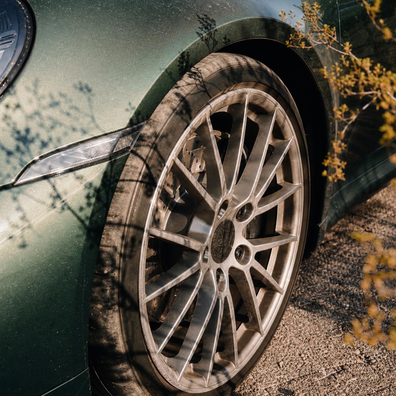 A tight detail of the front wheel and fender of an Aventurine Green Porsche 911, fine desert dust visible across the paint, the hard tactile shadow of a creosote branch falling diagonally across the metal