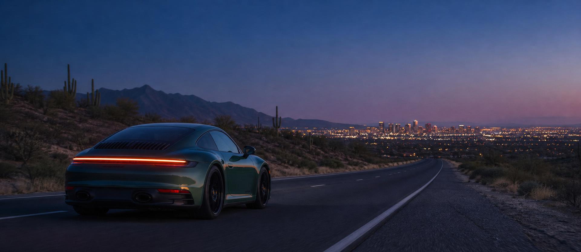An Aventurine Green Porsche 911 from behind driving south on an empty desert highway at deep blue hour, taillights glowing warm amber, the scattered warm lights of the Scottsdale skyline visible on the horizon, the McDowell Mountains silhouetted to the left
