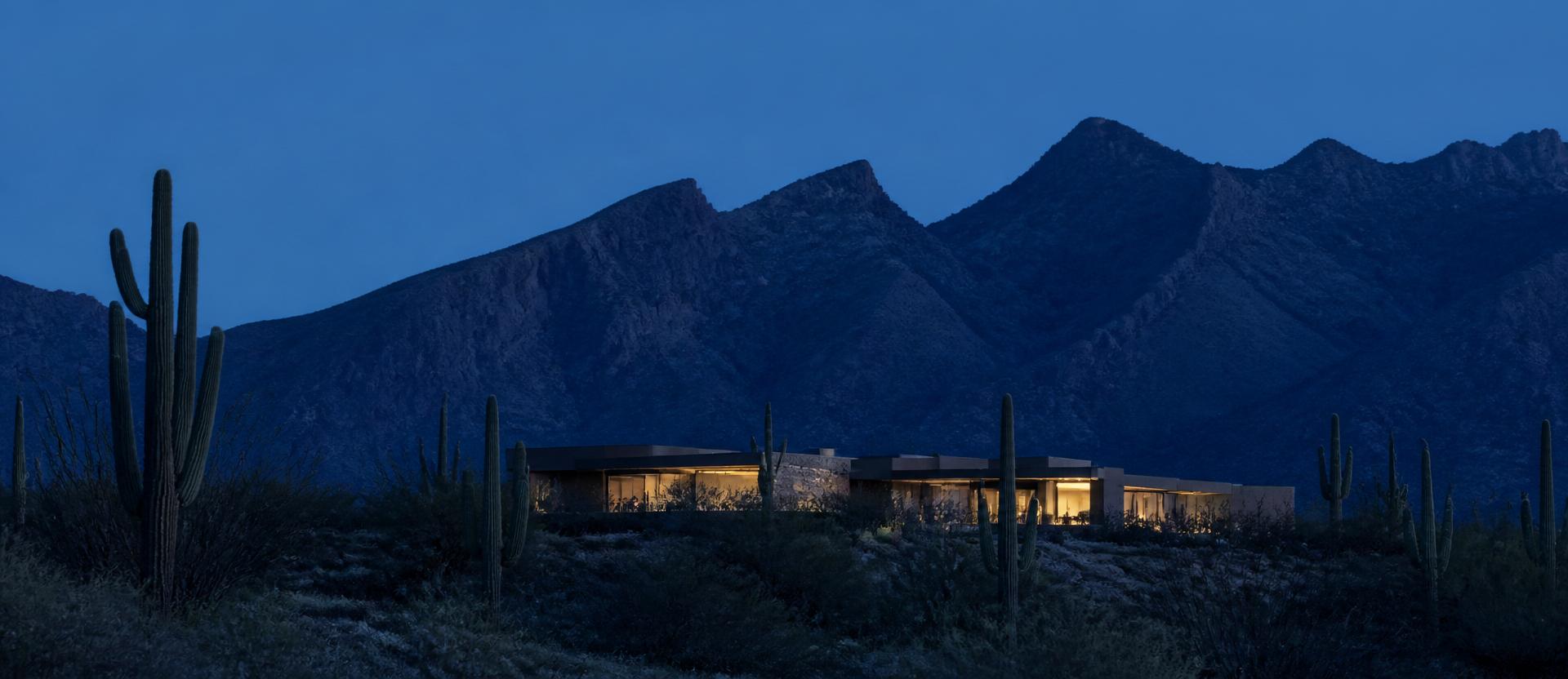 A wide blue-hour panorama of a low Sonoran modern Scottsdale residence in the middle distance with warm lamps glowing from within, set against the deep cobalt jagged silhouette of the McDowell Mountains, a scattering of saguaros across the foreground